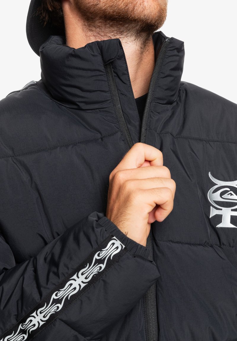 Czarna kurtka puchowa z wysokim kołnierzem; ma zamek błyskawiczny z przodu i szczegółowy tribalowy wzór na rękawie; gładki, lekki materiał.