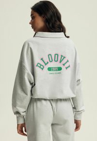 Szary sweter i pasujące spodnie z kolorową grafiką na plecach, która brzmi "BLOOVII 1989 CAMPUS DREAMER". Luźny krój i miękka struktura.
