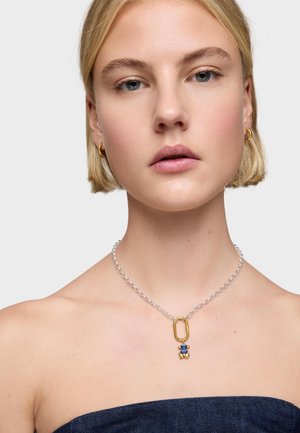 Junge Frau trägt kleine goldene Creolen und eine silberne Halskette mit einem Anhänger in Form eines Teddybären aus Gold, Blau und Perlen, vor einem schlichten Hintergrund.