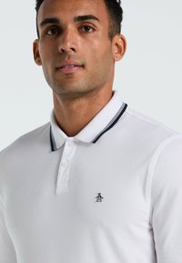 Witte polo shirt met zwart-wit gestreepte kraag, voorzien van drie knopen en een klein pinguïnlogo op de borst. Gladde textuur.