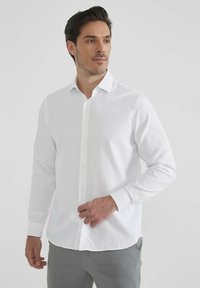 Chemise blanche à manches longues avec un col classique, fabriquée en tissu lisse, présentant un ourlet droit et des poignets buttonnés.
