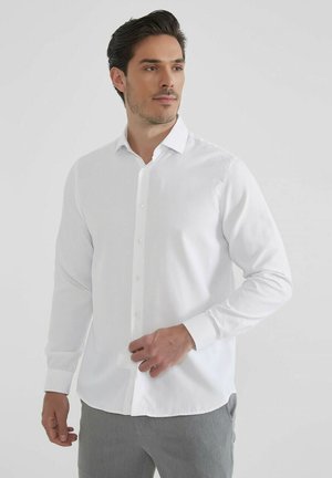 Chemise blanche à manches longues avec un col classique, fabriquée en tissu lisse, présentant un ourlet droit et des poignets buttonnés.