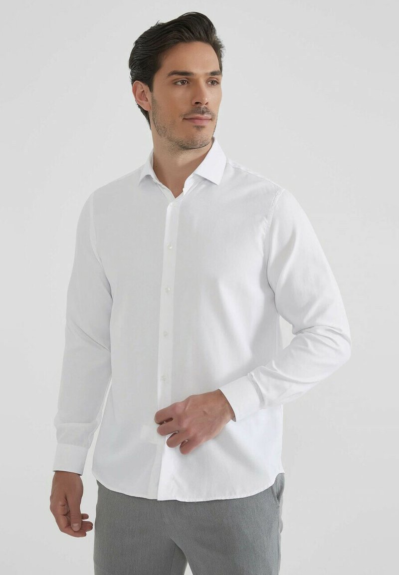 Chemise blanche à manches longues avec un col classique, fabriquée en tissu lisse, présentant un ourlet droit et des poignets buttonnés.