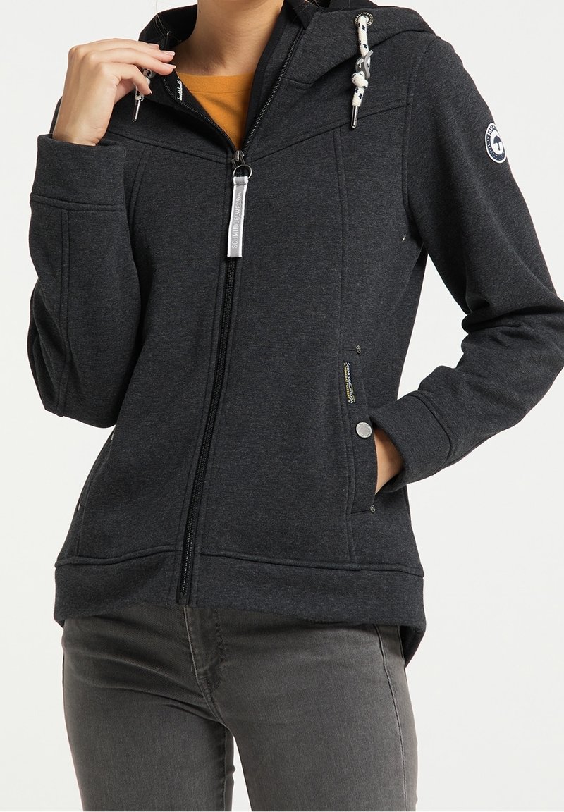 Femme portant un sweat à capuche zippé gris foncé avec cordons et poches latérales, associée à un pantalon gris et un t-shirt orange en dessous.