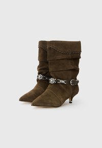 Stivaletti in suede marrone al polpaccio con design morbido, punta appuntita e dettagli in acciaio inox. Presentano una rifinitura intrecciata lungo la parte superiore.