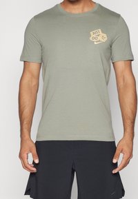 Camiseta de algodón verde oliva con cuello redondo y mangas cortas. Presenta un logo dorado de "Just Do It" en la parte superior izquierda del pecho.