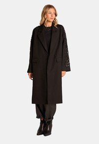 Abrigo de lana negro con mangas largas que presenta un intrincado bordado negro. Solapas con muesca y una silueta relajada. Combinado con pantalones ajustados negros.