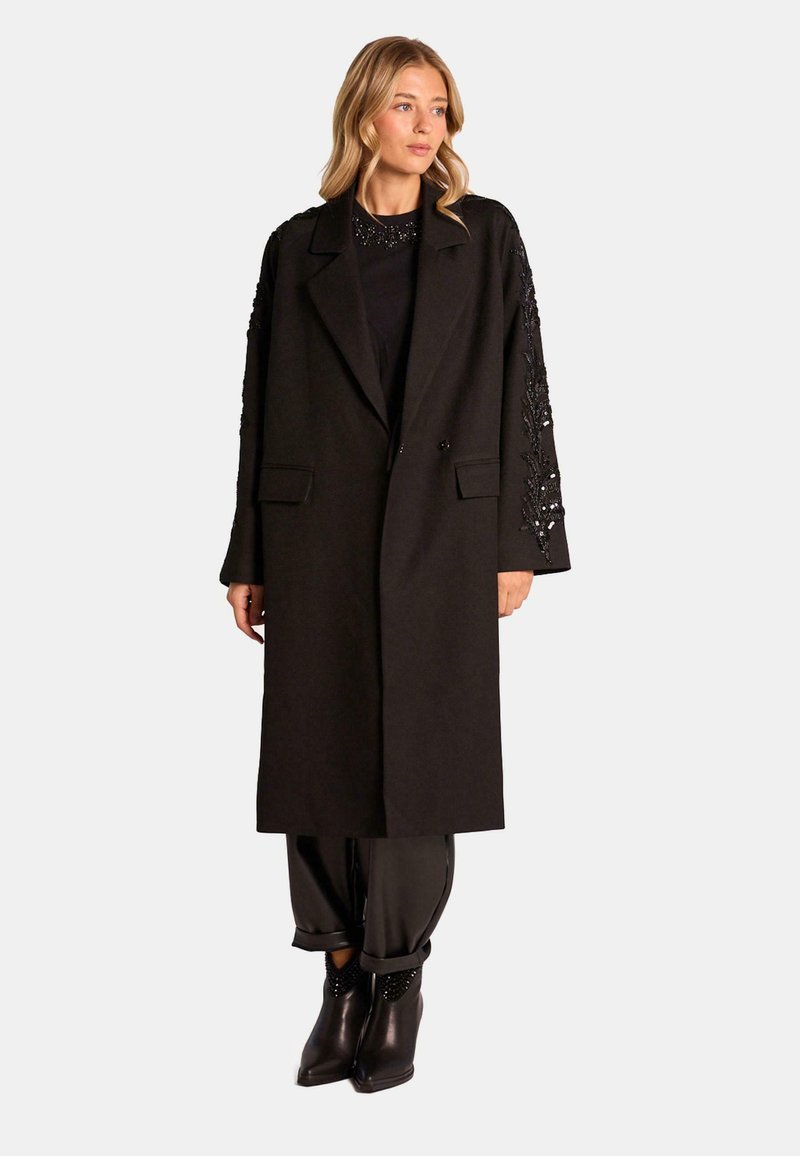 Abrigo de lana negro con mangas largas que presenta un intrincado bordado negro. Solapas con muesca y una silueta relajada. Combinado con pantalones ajustados negros.