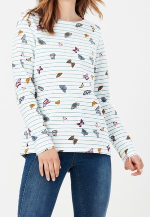 Kvinde iført en hvid langærmet skjorte med lyseblå striber og farverige sommerfugleprint, kombineret med mørkeblå jeans.