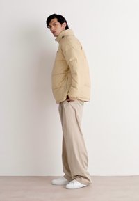 Beige puffer jacket with a high collar, quilted texture, and black zipper, paired with loose beige trousers and white sneakers.