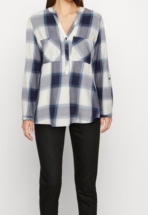 Femme portant une chemise à carreaux bleue et blanche à manches longues avec deux poches avant et un pantalon noir, debout devant un fond uni.