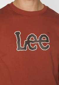 Červená bavlněná mikina s výrazným černobílým logem "Lee" v hravém písmu, s kulatým výstřihem a žebrovaným límcem.