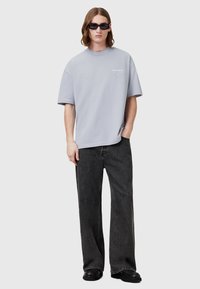 Tricou oversized gri deschis cu guler rotund și imprimeu logo, asortat cu blugi negri largi și încălțăminte neagră.