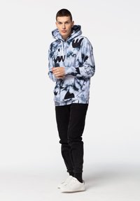 Sudadera con capucha en azul tie-dye con patrones abstractos en negro. Combinada con pantalones negros y zapatillas blancas. Ajuste casual y tela texturizada.