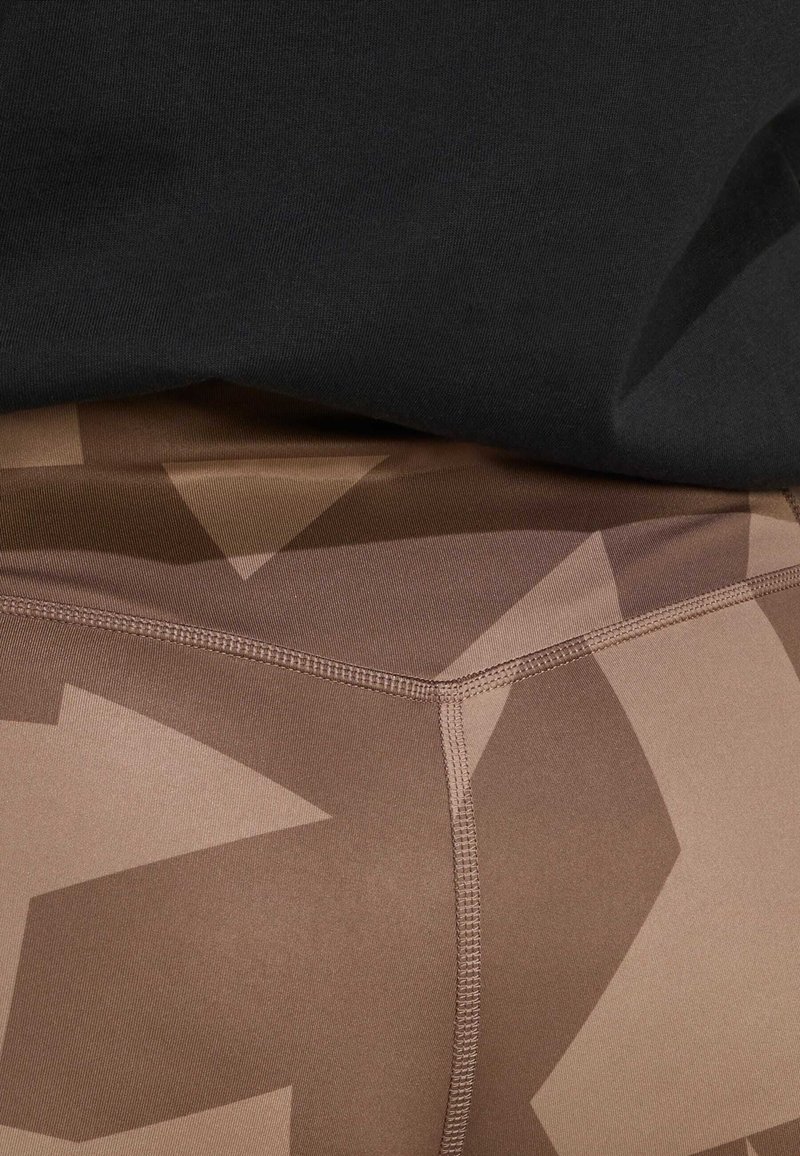 Leggings a motivo camouflage marrone con una texture liscia, caratterizzati da motivi angolari. L'elastico in vita è parzialmente coperto da un top nero.