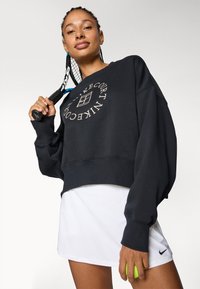 Felpa nera corta con design a logo rotondo, abbinata a una gonna da tennis bianca. Il modello tiene in mano una racchetta da tennis e una pallina.