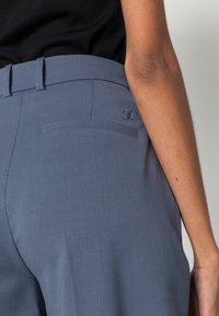 Zbliżenie na osobę w niebieskich, dopasowanych spodniach z tylną kieszenią i wyszywanymi inicjałami, w połączeniu z czarną bluzką.