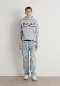 Sweat à capuche gris avec le logo GANT 1949, associé à un jean délavé bleu clair. Le modèle porte des baskets blanches. Tenue simple et décontractée sur un fond uni.