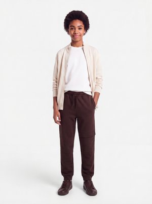 Ung person med kort krøllet hår iført en hvid skjorte, beige jakke, mørkebrune bukser og mørkebrune sneakers, står med den ene hånd i lommen.