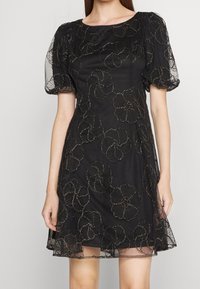 Robe noire avec des manches courtes bouffantes, design floral brodé, superposition transparente et silhouette ajustée, avec des accents métalliques tout au long.