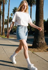 Ung kvinde går på en solrig fortov omgivet af palmer, iført en hvid t-shirt, denimskørt, hvide sokker og hvide sneakers.