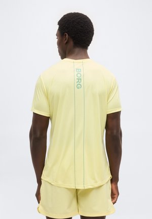 Muškarac u svijetložutoj sportskoj odjeći s natpisom „BORG“ napisanim okomito na stražnjoj strani majice, stoji ispred jednobojne pozadine.