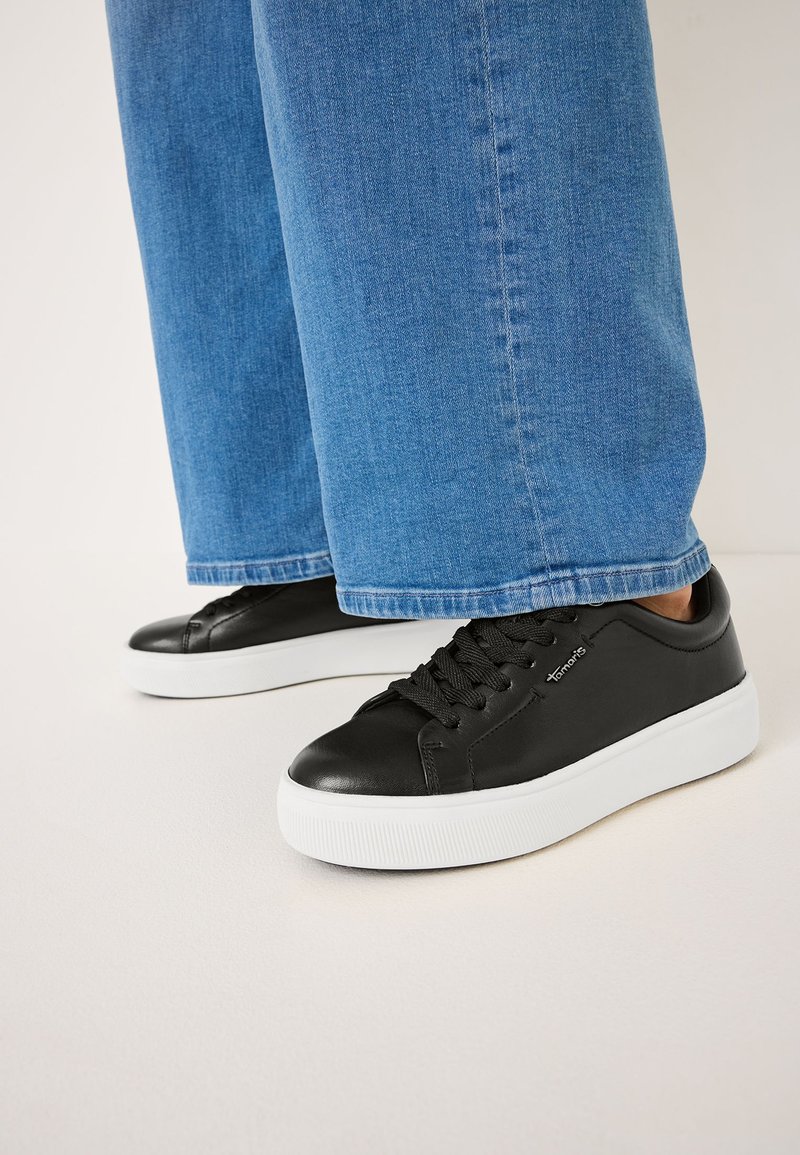 Sneaker in pelle nera con suole in gomma bianca e lacci neri, mostrati insieme a pantaloni in denim blu con vestibilità rilassata e orli risvoltati.