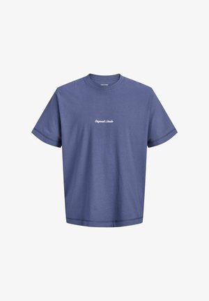 Teenage dreng med krøllet hår står smilende i en blå T-shirt, blå jeans og beige sko mod en ensfarvet lys baggrund.