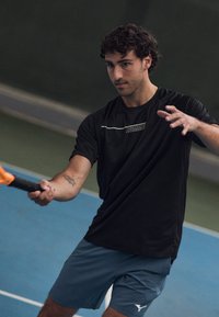 Mann med krøllete hår i svart skjorte og blå shorts spiller paddle tennis innendørs, holder racket og fokuserer på ballen.