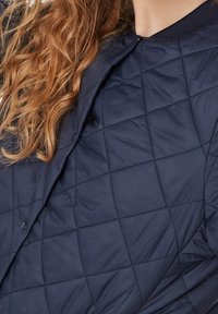 Marinfärgad kviltad jacka med diamantmönster, tryckknappsstängning och ribbad krage. Materialet verkar lätt och texturerat.