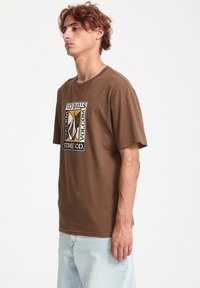 Brun bomulds t-shirt med et centralt grafisk motiv i sort og gul, korte ærmer og en afslappet pasform, parret med lyseblå jeans.