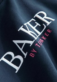 Jūras spēku džemperis ar uzkrītošu baltu un rozā izšūtu tekstu "BAKER by TEVAKE" un mīkstu, teksturētu audumu.