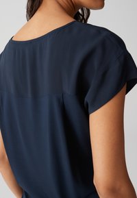 Top blu navy con una texture liscia, maniche corte e un ampio scollo che presenta un sottile dettaglio di cucitura sul retro.