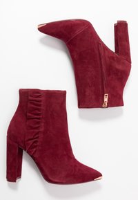 Stivali ankle in suede bordeaux con punta appuntita, tacco alto a blocco e zip laterale. Presentano dettagli arricciati su un lato e un accento metallico.