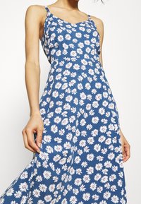 Robe fleurie bleue avec des bretelles spaghetti, présentant un motif de marguerites en blanc et rose, fabriquée en tissu léger, fluide et légèrement cintrée.