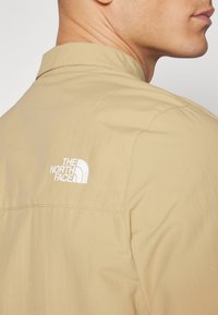 Beżowa lekka koszula z kołnierzykiem, z białym logo na górnej części pleców oraz subtelną fakturą. Brak wyraźnych wzorów lub akcentów.