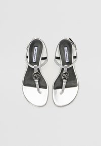 Sølvmetalliske sandaler med en thong-rem, justerbar ankelspenne og en sirkulær logoaccent. Svart såle med en jevn overflate.