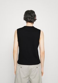 Person mit kurzen dunklen Haaren, trägt ein ärmelloses schwarzes Oberteil und eine hellgraue Hose, von hinten vor einem schlichten weißen Hintergrund.