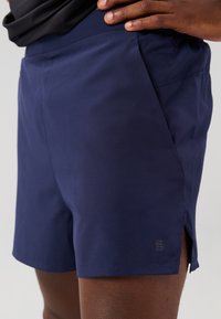 Navy blue athletic shorts with a smooth texture, side slits for mobility, and a small logo near the hem. Lightweight and flexible fabric.