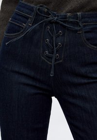 Σκούρο μπλε τζιν από denim με δέσιμο μπροστά σε φιόγκο, ορατές τσέπες και πορτοκαλί ραφές.