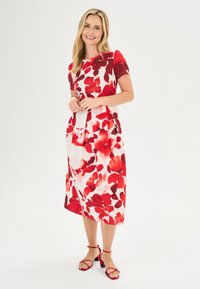 Rotes und weißes Blumenkleid mit kurzen Ärmeln, das einen plissierten Rock hat. Das Model trägt rote Sandalen mit Riemen vor einem neutralen Hintergrund.