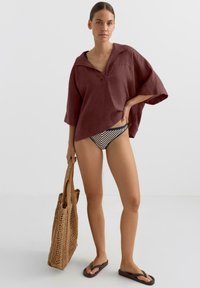 Bordó len blúz V-nyakkal és széles ujjakkal, csíkos bikini alsóval, barna papucsokkal és fonott bevásárlótáskával párosítva.