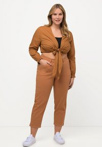 Brun, kortærmet bluse med knude foran og lange ærmer, parret med matchende brune tapered bukser og hvide sneakers.