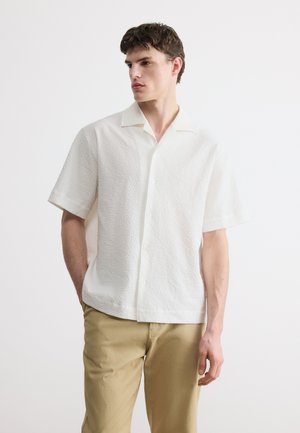 Jeune homme aux cheveux bruns courts portant une chemise blanche texturée à manches courtes avec des boutons et un pantalon beige, debout devant un fond uni.