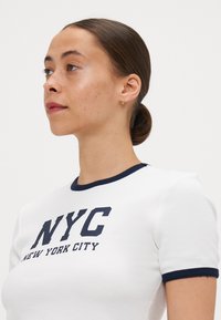Hvit tettsittende t-skjorte med marineblå kant, med stort "NYC" trykk og "NEW YORK CITY" tekst i kontrastfarge blå. Glatt tekstur, korte ermer.