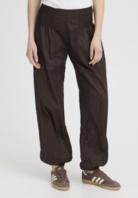 Personne portant un pantalon noir ample avec taille et poignets élastiques, associé à des baskets Adidas marron avec des bandes blanches.