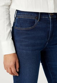 Diepblauwe denim jeans met een slim fit, afgewerkt met contrasterende gele stiksels, voorzakken en een subtiele textuur. Vergezeld van een wit shirt.