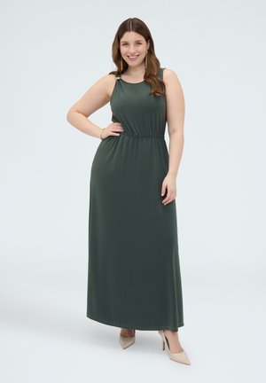 Donna sorridente con lunghi capelli castani che indossa un abito lungo verde scuro senza maniche e tacchi beige, in piedi con la mano sui fianchi su uno sfondo uniforme.