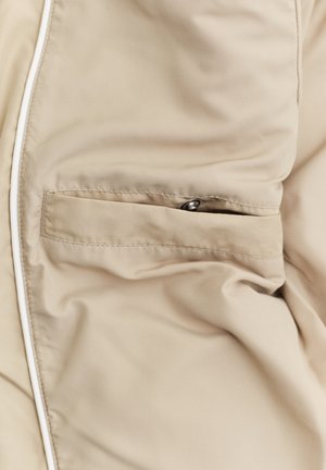 Beżowa tkanina z przeszytą poziomą kieszenią z małym metalowym zatrzaskiem i białą lamówką wzdłuż krawędzi.