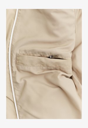 Beżowa tkanina z przeszytą poziomą kieszenią z małym metalowym zatrzaskiem i białą lamówką wzdłuż krawędzi.